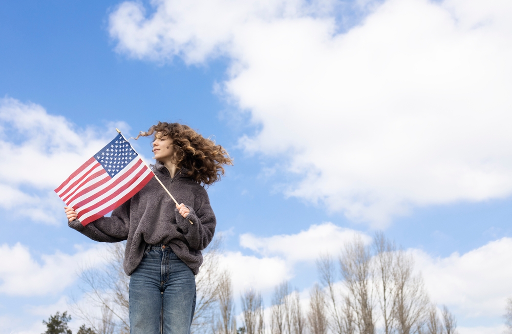 A young girl holding an American flag with cloudy blue skies overhead