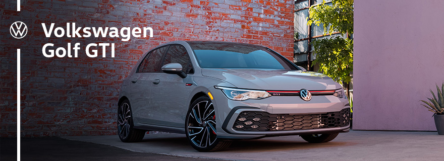 Front passenger side view of a grey 2025 Volkswagen Golf GTI parked on a driveway with a brick wall behind on a sunny day